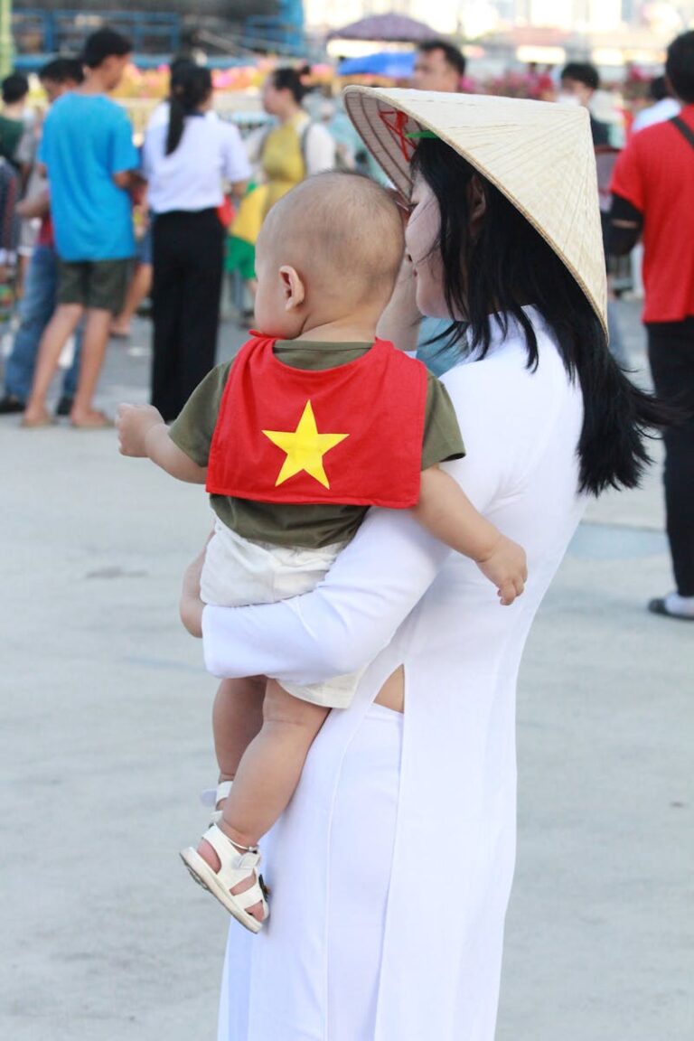 A woman in traditional dress holds her child during a vibrant outdoor parade in Vietnam.